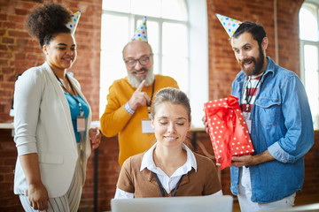Young manager networking in front of laptop while her colleagues preparing her birthday surprise behind