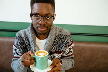 Handsome pensive afro american man in eyeglasses and sweater having black coffee latte during lunch, sitting alone at cafe, having thoughtful look, looking at camera. Warming in snow winter day.