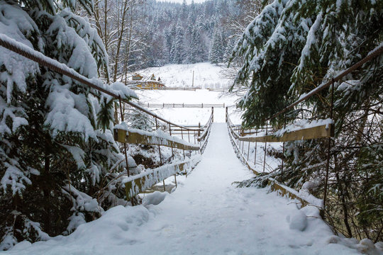 Fototapeta old suspension bridge across river, winter fir forest in snow