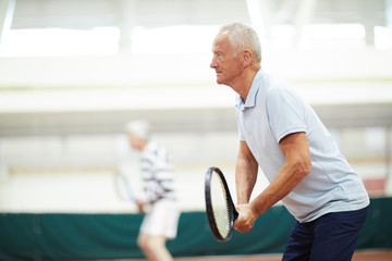 Active mature tennis player with racket and his mate on background concentrating on game