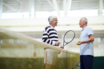 Two happy senior tennis players with equipment standing by net on court and discussing some moments...