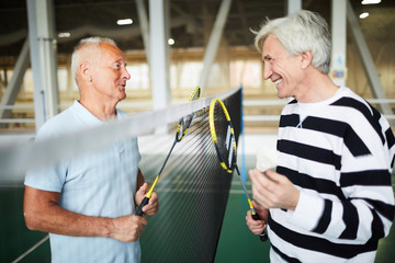 Two friendly senior badminton players in activewear with rackets standing by net and interacting