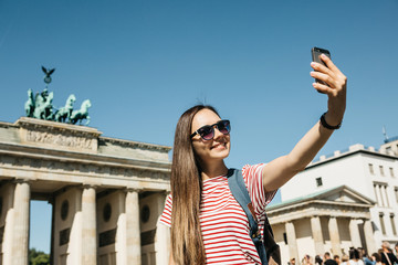 Young beautiful positive girl makes selfie against the background of the Brandenburg Gate in Berlin in Germany or takes pictures of sights.