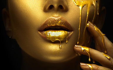 Golden paint smudges drips from the face lips and hand, golden liquid drops on beautiful model girl's mouth, creative abstract makeup. Beauty woman face - obrazy, fototapety, plakaty