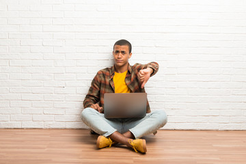 African american man sitting on the floor with his laptop showing thumb down sign with negative...