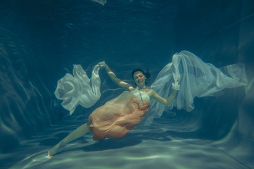 elegant slender girl swims underwater like a free diver in a white evening dress with beautiful...