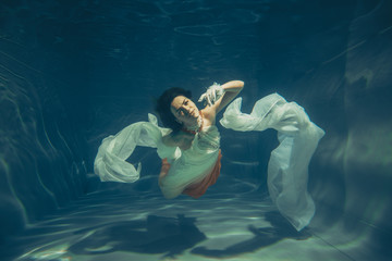 elegant slender girl swims underwater like a free diver in a white evening dress with beautiful...
