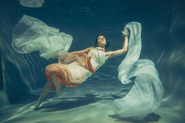 elegant slender girl swims underwater like a free diver in a white evening dress with beautiful...