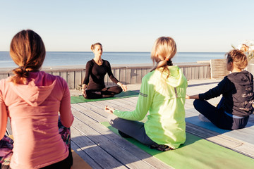 Fitness, people and healthy lifestyle concept - group of women meditating in lotus pose outdoor on wooden terrace. Yoga class with teacher in nature by the sea against blue sky background, copyspace
