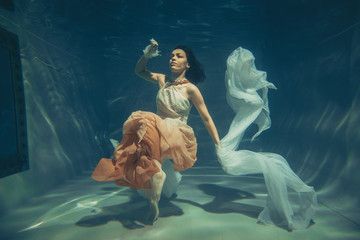 elegant slender girl swims underwater like a free diver in a white evening dress with beautiful...