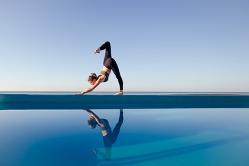 Young attractive woman practicing yoga stand doing Downward facing dog exercise, adho mukha svanasana pose, working out by the pool, above beach, relaxing against blue sky. Health and beauty concept