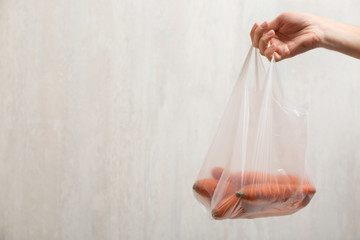 Woman holding plastic bag with carrot on light background, closeup. Space for text