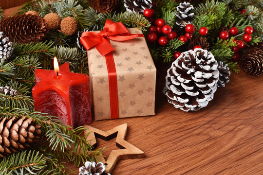 Gft and candle as part of Christmas decoration made of fir branches, holly and pinecones on wooden desk