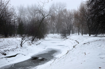 Small river with melting snow and ice in cloudy foggy weather in the city park