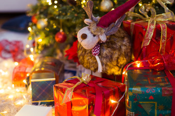 Toy deer between presents and christmas tree on the background. Red and green boxes with different patterns and golden tapes. Blur lights.