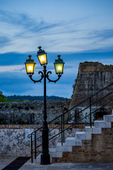 Vintage illuminated street lamp at dawn with three lights in front of ancient white stone steps of staircase found in the medieval fortress in Corfu Island, Kerkira, Greece. Springtime cloudy evening