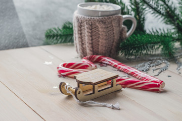 White cup for cocoa in beige knitted clothes. Santa Claus sleigh on a white wooden background with christmas tree and red caramel candy
