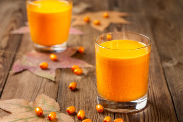 Two glasses of fresh pressed sea buckthorn juice, with autumn leaves and sea buckthorn berries on a wooden table. Blurred background.