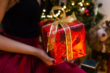 Hands are holding present in the red box with pattern and golden tape around. Women in evening dress. Blur lights on the background