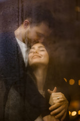 Fototapeta na wymiar Loving guy hugs and kisses his happy girlfriend standing behind a wet window with lights. Romantic couple
