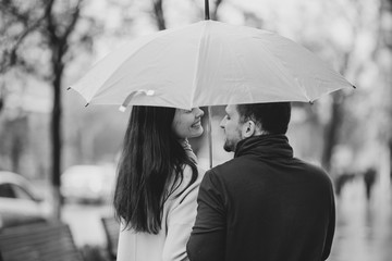 Happy beautiful couple, guy and his girlfriend dressed in casual clothes stand under the umbrella and look at each other on the street in the rain. Black and white photo