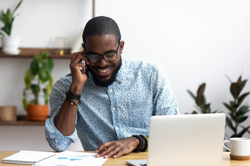 Handsome african manager sitting at office desk in front of laptop hold mobile phone make pleasant business or informal call. Successful businessman looking at financial statistic shown on documents - Powered by Adobe