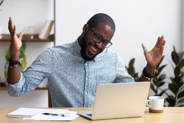 Joyful black businessman sitting at desk looking at computer screen talking with friend make...