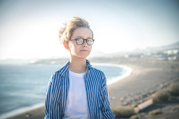 Blond child with eyeglasses and white striped blue shirt smiles in the wind, freedom, joy, happiness, life, beach in the background, sea and blue sky