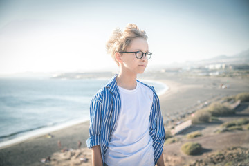 Blond child with eyeglasses and white striped blue shirt smiles in the wind, freedom, joy, happiness, life, beach in the background, sea and blue sky