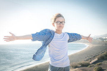 Blond child with eyeglasses and white striped blue shirt smiles in the wind with arms wide open, freedom, joy, happiness, life, beach in the background, sea and blue sky