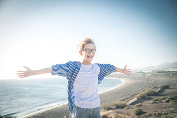 Blond child with eyeglasses and white striped blue shirt laughs in the wind with arms wide open, freedom, joy, happiness, life, beach in the background, sea and blue sky
