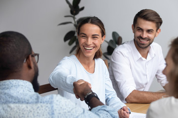 Attractive young mixed race woman shaking hands with black partner starting negotiations sitting together in office boardroom with colleagues company members millennial staff feels good and satisfied. - obrazy, fototapety, plakaty