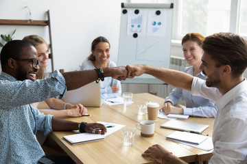 Diverse company staff girls guys sitting at desk in boardroom feel happy and satisfied celebrating success at work. Diverse colleagues fist bumping greeting each other express friendship and respect