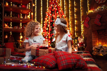 Happy little girls wearing Christmas pajamas open gift box by a fireplace in a cozy dark living room on Christmas eve. Celebrating Xmas at home.