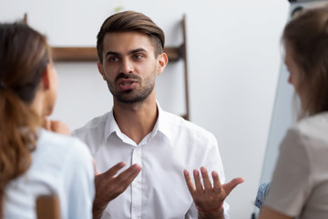 Millennial confident male worker express his opinion about business plan share thoughts with rear view female colleagues. Candidate for job vacancy answering on HR managers questions during interview