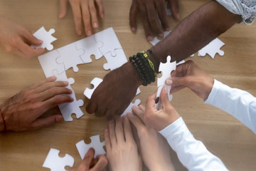 Top above close up view diverse multinational businesspeople hands assembling together puzzle scattered on table. Symbol of teamwork and partnership, business strategy and logic thinking concept