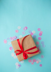 Gift box wrapped in brown craft paper and tied with red bow on blue background with colofrul confetti and copyspace.