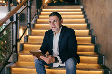 Smiling middle-aged Caucasian businessman dressed smart casual sitting on the stairs in cafe while...