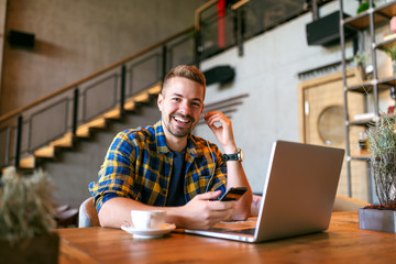 Portrait of young smiling Caucasian freelancer holding smart phone and putting on earphones while sitting in cafe. On the desk laptop and coffee. - obrazy, fototapety, plakaty