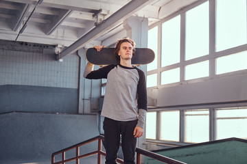 Young skateboarder standing next to a grind rail in skatepark indoors,  holding his board and looking away.