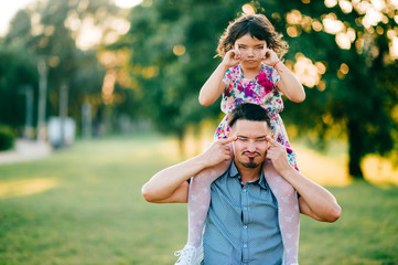 Obraz premium Lifestyle sunset portrait of unusual couple of dad with daughter on shoulders making funny expressive faces. Happy family playing at summer nature outdoor. Fatherhood and childhood. Dad loves his girl