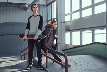 Attractive youth couple with skateboards next to a grind rail in skatepark indoors.