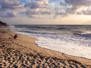 Man with smartphone photographs a wave crashing on the beach at sunset.