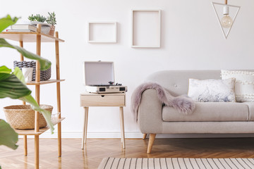 Stylish and cozy scandinavian white interior with design sofa, bookstand with accessories, pillows, blanket, gramophone and mock up photo frames. White background walls and modern triangle lamp. 
