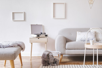 Stylish and cozy scandinavian white interior with design sofa, bench, coffee table, pillows, blanket, gramophone and mock up photo frames. White background walls and modern triangle lamp. 