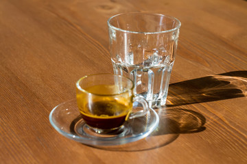 A transparent cup of coffee ground and an empty  glass stand on the table. The morning sun shines on the cup and glass, their shadows on the table.