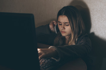 Sad teenage girl sitting near laptop in dark room. She is a victim of online bullying Stalker...