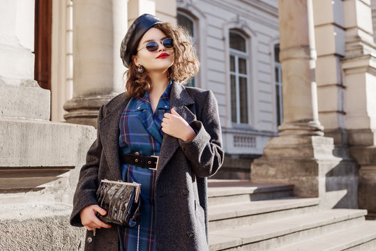 Outdoor fashion portrait of young beautiful fashionable girl wearing blue checkered dress, grey coat, leather beret, sunglasses, holding small bag, posing in european city. Copy, empty space