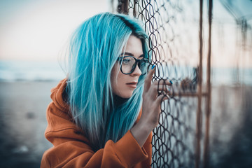 Portrait of young hipster woman with blue hair and glasses sitting on beach near mesh fence - Powered by Adobe