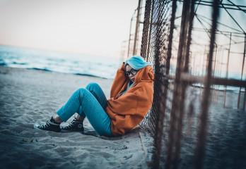 Young hipster woman with blue hair and glasses sitting on beach near mesh fence